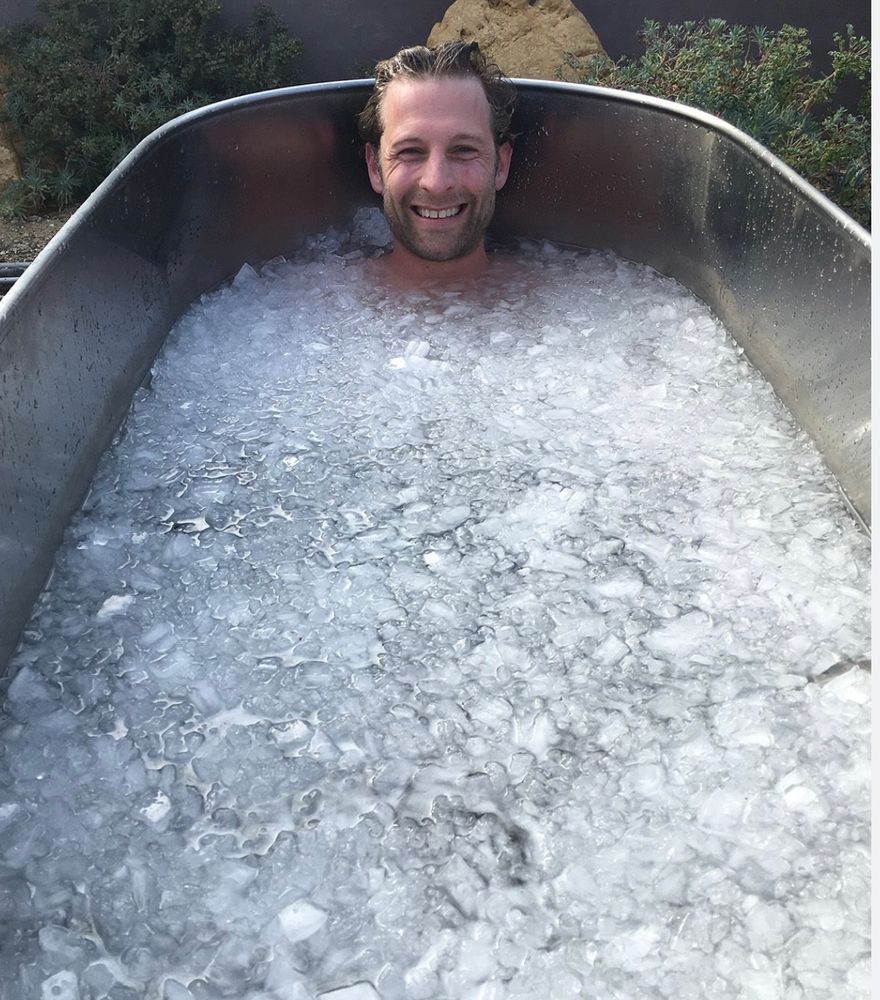 A young man’s smiling face sticking out of a huge bathtub full of ice.