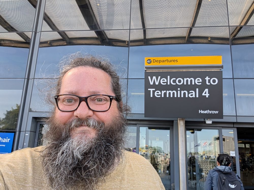 A very hairy man stands in front of a sign at Heathrow Airport reading: Departures. Welcome to Terminal 4.