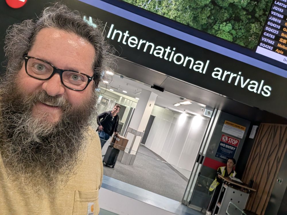 A very hairy man stands in front of a sign in Auckland International Airport which reads: International Arrivals