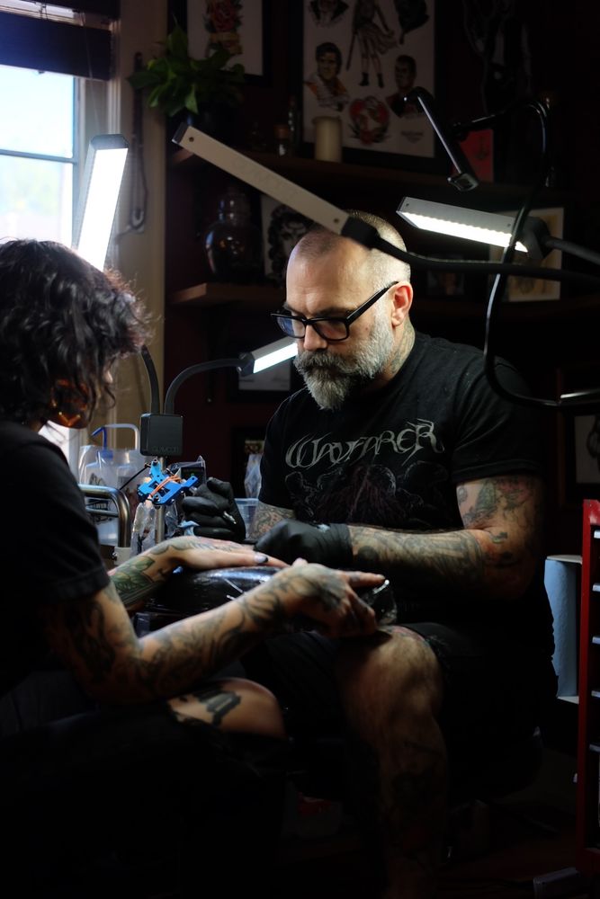 A male tattoo artist with a shaved head and a graying beard, wearing a black band shirt, tattoos his clients knuckles