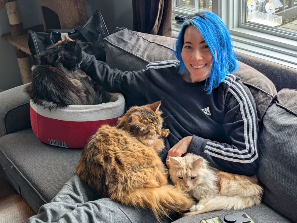 Erika, a person with short blue hair, sits on the couch with 3 of their 4 cats.