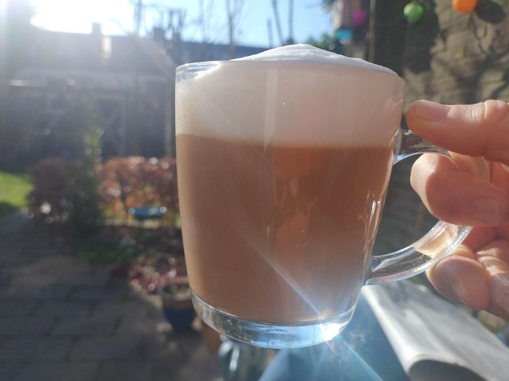 Zonlicht schijnt op vastgehouden glas koffie met melkschuimlaag, tuin en huizenrij op achtergrond. 