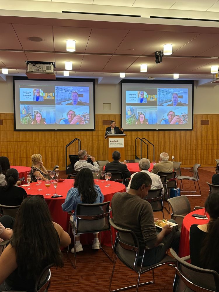 A group of colleagues seated, listening to Prof. David Grusky at a podium speaking about Prof. Mark Granovetter. A slide of Mark Granovetter and people attending on Zoom are projected behind the podium.
