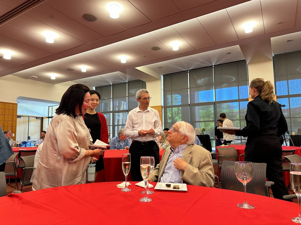 Prof. Mark Granovetter, seated, with champagne and brownies, speaking to a few colleagues standing next to him.