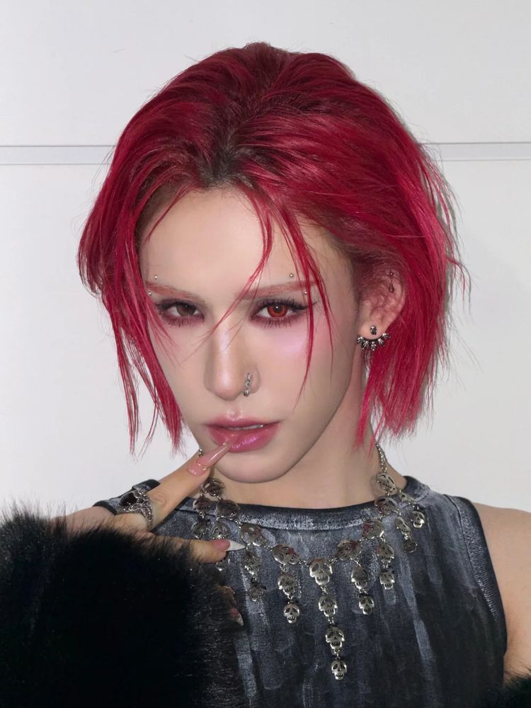 xlov wumuti with red slicked hair with some locks in her face. her eyelashes are red too with eyebrow piercings. she has a nose pin on and pink lips. she is wearing a grey tank top with a necklace of skulls and has her acrylic nail on her lower lip while smizing