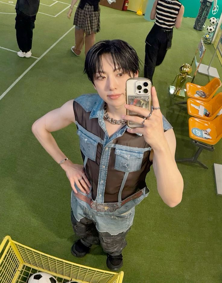 andteam fuma at an indoor soccer field wearing a denim and mesh vest and jeans with one arm bent at the elbow with his hand on his hip and the hand being used for taking the selfie