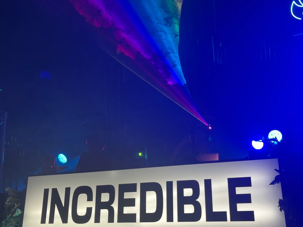 The word “Incredible” on a backlit sign, below a rainbow swoop of laser light and smoke 