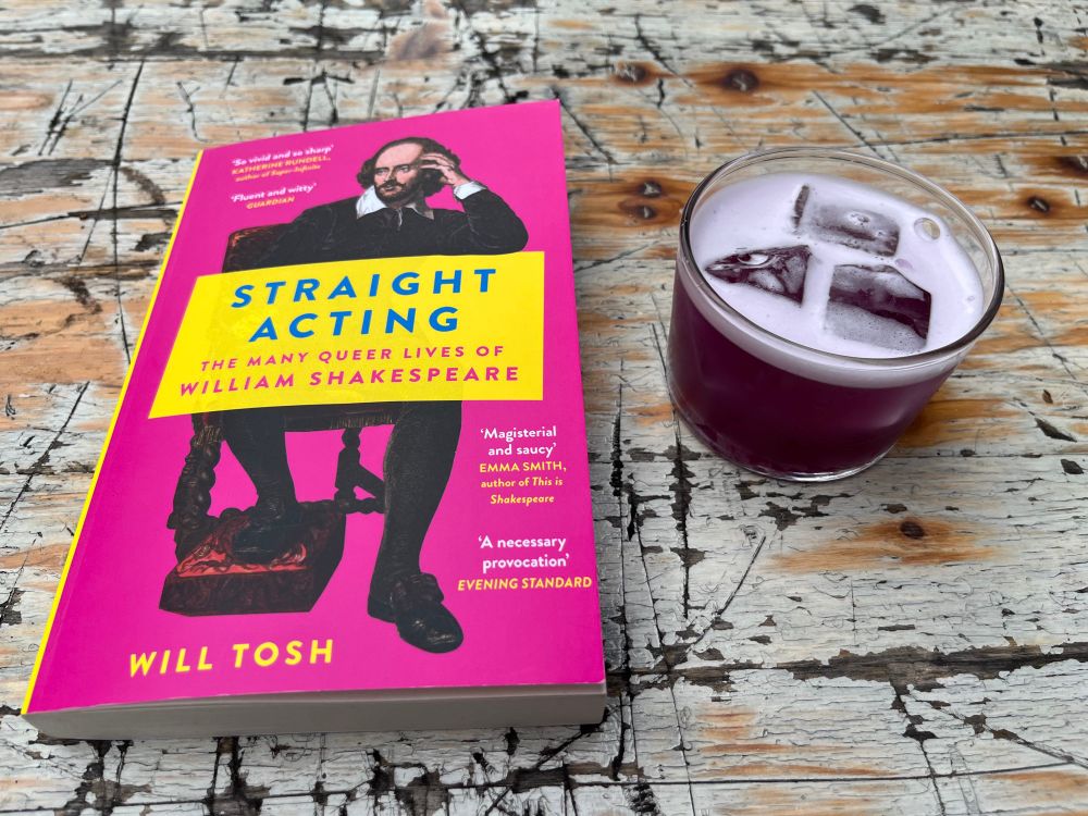 Book with a bright pink cover and purple drink on a scratch up table. Shakespeare is posing on the cover as if he’s a hipster in an indie band. The title reads Straight Acting, the many queer lives of William Shakespeare by Will Tosh. To the right is a cute lil tumbler of a purple cocktail with 3 big chonks of ice, it tastes like slowly unwinding after a long week. 