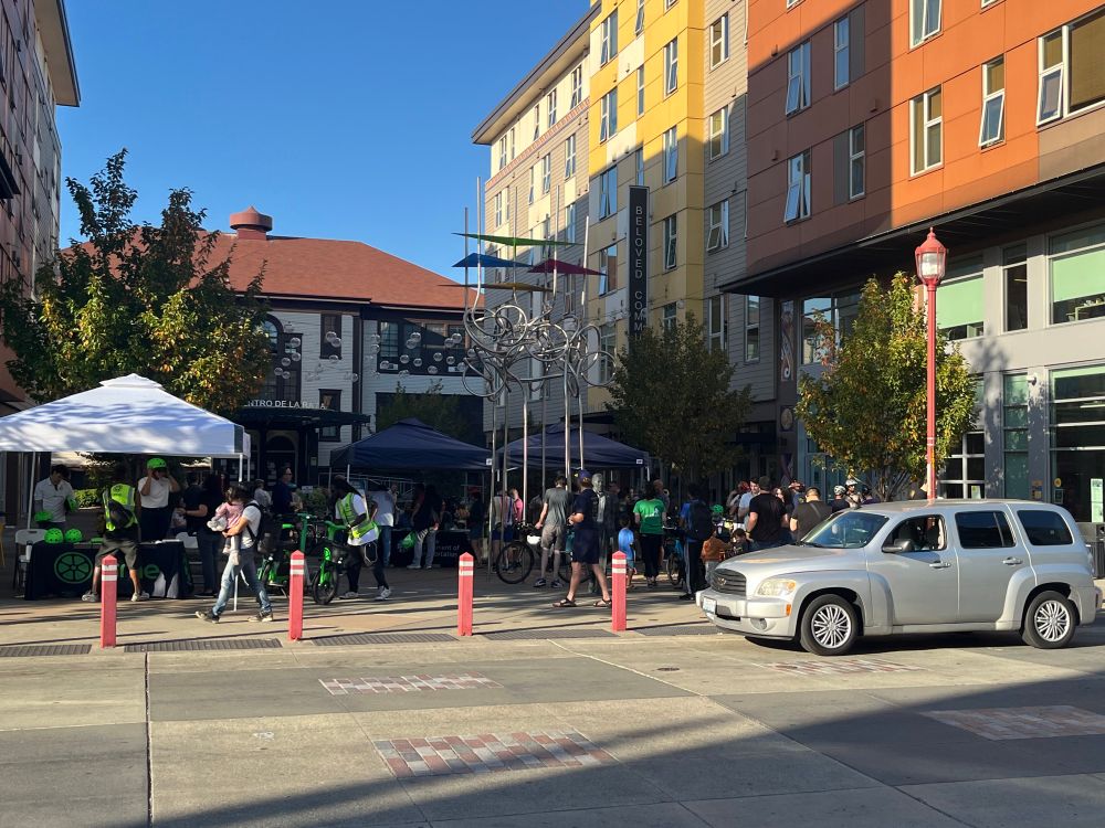 About 30 people near three booths. A car is parked in front of the celebration. 