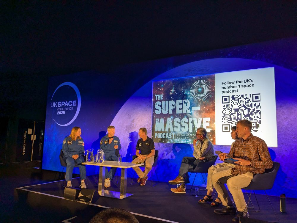 Panel sitting, Meganne Christian and John McFall in blue jumpsuits, Tim Peake with a dark blue shirt and off white pants, to the left sitting the podcast host and producers. Background with an close up of the Moon and conference logo and the super massive podcast logo