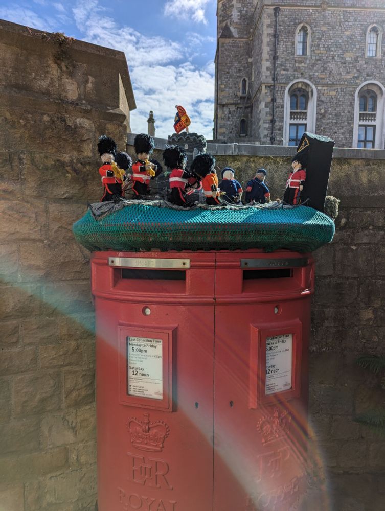 Red postbox double size , two mail slots letters of Elizabeth 2nd R and Royal mail. Crochet top of royal guards on parade with different musical instruments. Clear blue skies and a small part of Windsor castle in the background 