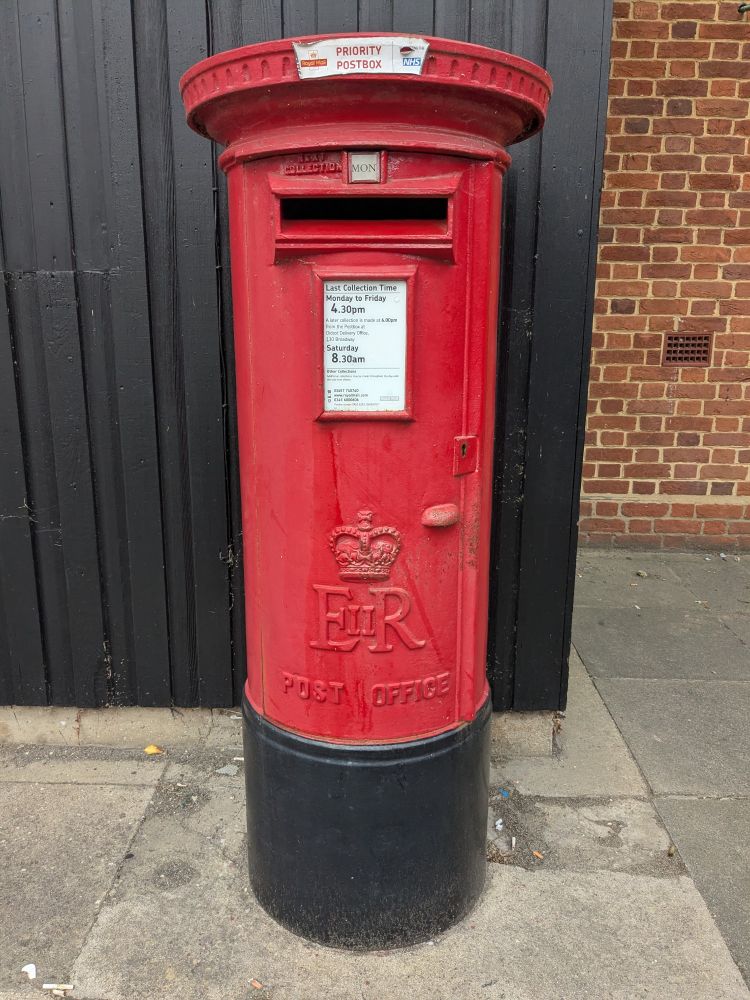 Red postbox, circular round top and cylinder long middle, bottom is black. Post office times written in a white rectangle box in the middle. Underneath a crown with the letters E II R (the II part much smaller between the E and the R for Elizabeth 2nd Regina) and the words post office. Background of brown bricks and black fance