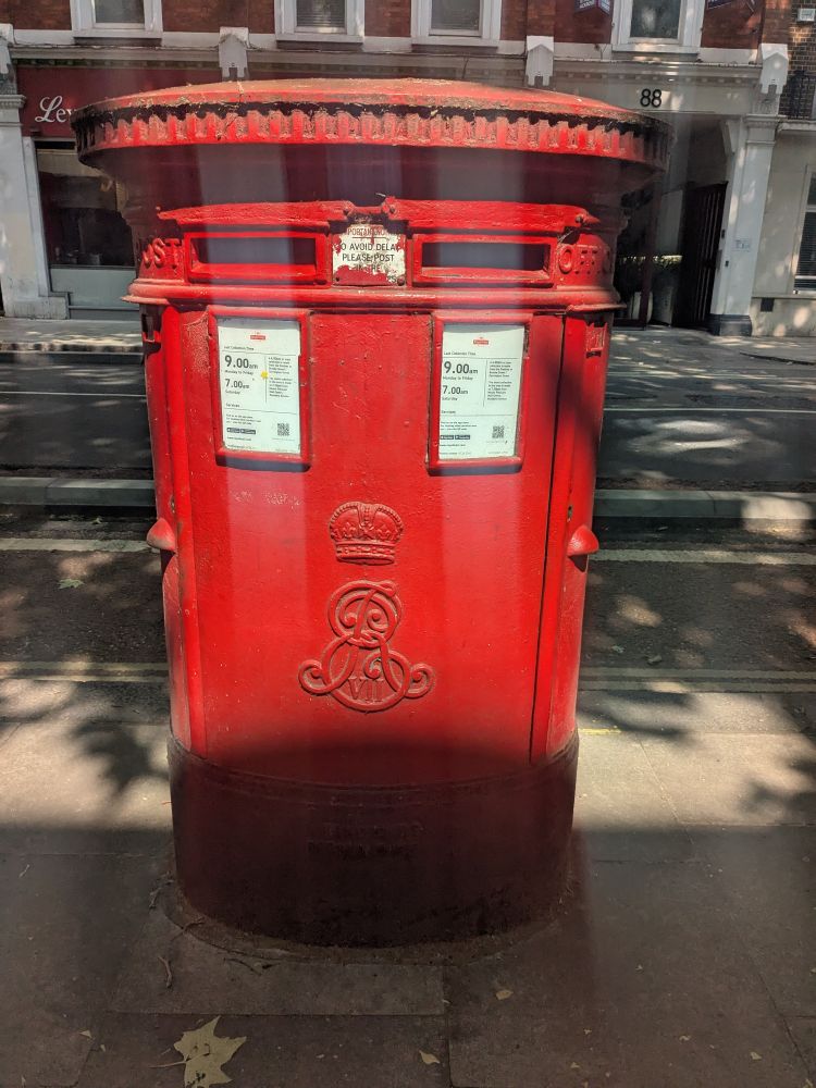A big red postbox, double in size compared to others with two slots on top to put mail inside. Big round top and massive body, crown of Edward the 7th with the letters E and R entwined and the letters vii very small inside the E. Background of a London street