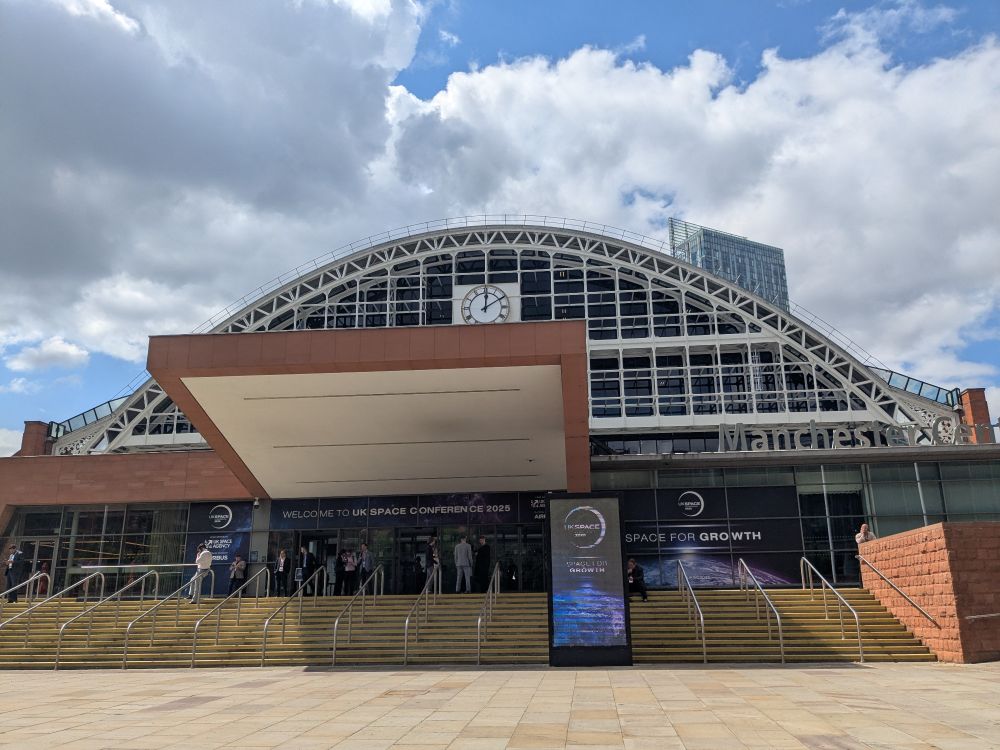 The front of the Manchester conference centre with big signs of the UK space conference. Conference center has a half Dome shape at the front. Clear blue skies with white clouds.