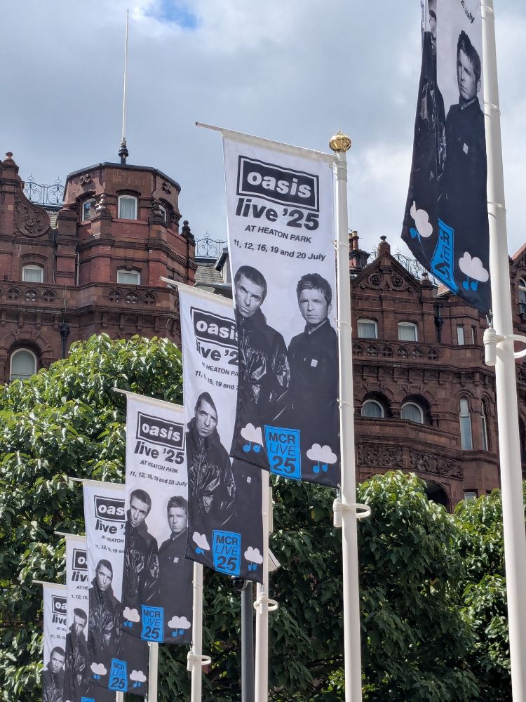 Line of flags for the Oasis live tour, showing Oasis and saying live 25, background of some trees and red brick tall houses 