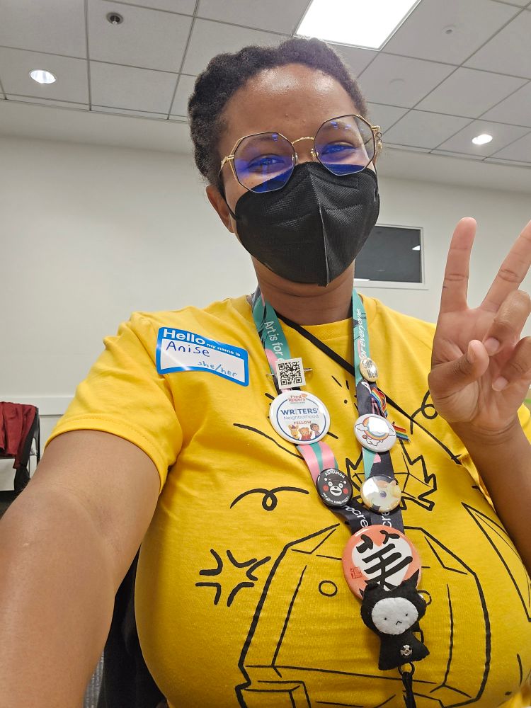 Anise giving a peace sign, wearing her yellow LightBox Expo Firefly shirt and lanyard adorned with pins