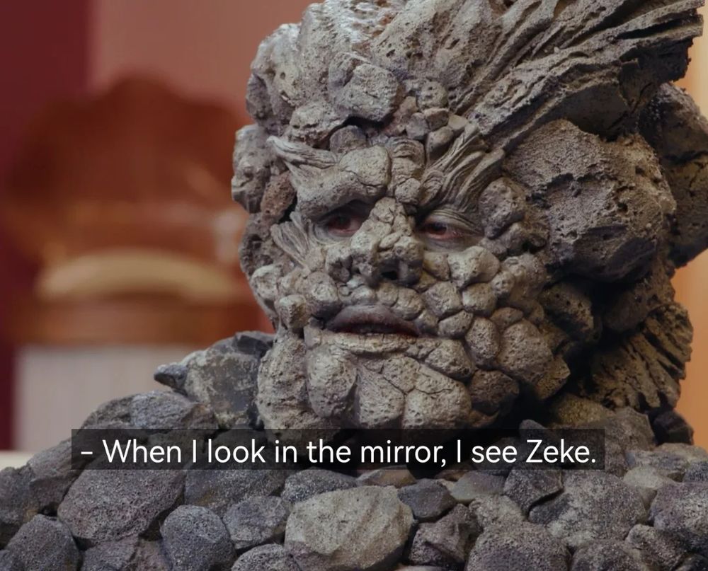 A picture of a person costumed to be made of rocks from Dropout's show Very Important People, saying "When I look in the mirror, I see Zeke" 