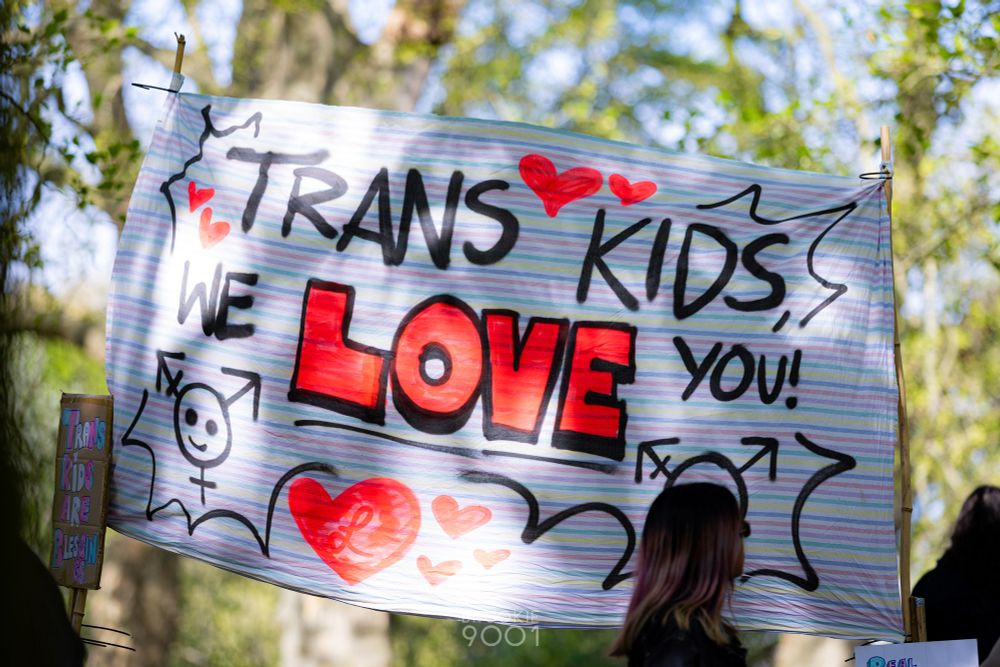 Sign reading "Trans Kids We Love You" with hearts and trans symbols drawn on it too