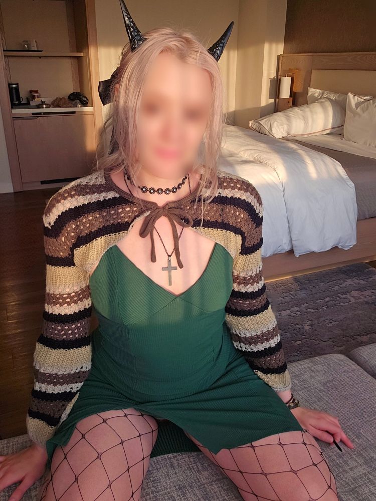 White woman in a tight green dress, that highlights her slim figure, wearing fishnets in front of mirror. She has horns, a cross neckless, and a chocker. Her hair is in a bow, which you can see a glimpse of. She has a striped green knit cardigan on that only covers her shoulders. Her dress rides up very close to danger zones.
