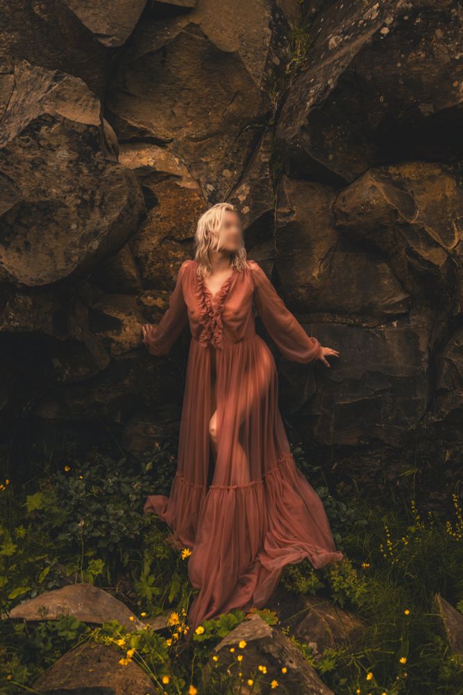 White women stand against a wall of wrongs, amidst greenery and yellow followers, wearing a dusty pink "kill your husband" gown. you can see some dark circles where the chest is, and the highlight of a leg through the sheer fabric. 