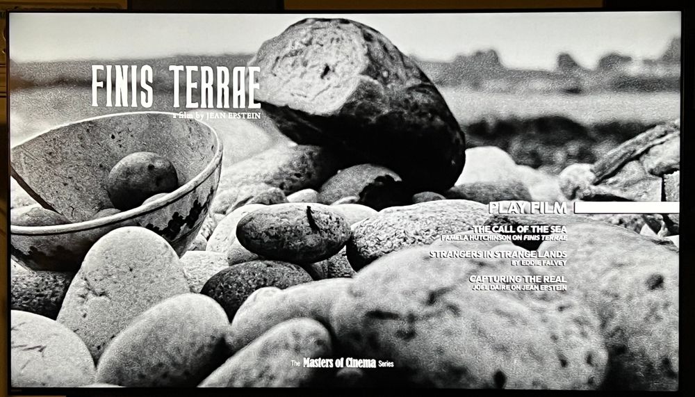 Image of the menu for the film FINIS TERRAE. The title of the film is in the top left and the three bonus features are listed in the bottom left. The collection title “The Masters of Cinema Series” is on the bottom of the screen. The rest of the screen is taken up by bread and a bowl resting on rocks.