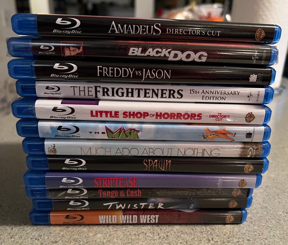 Stack of Blu-rays sit on a counter with the films in alpha order:
AMADEUS: Director’s Cut
BLACK DOG
FREDDY VS JASON
THE FRIGHTENEES
LITTLE SHOP OF HORRORS
THE MASK
MUCH ADO ABOUT NOTHING
SPAWN
STRIPTEASE
TANGO & CASH
TWISTEE
WILD WILD WEST