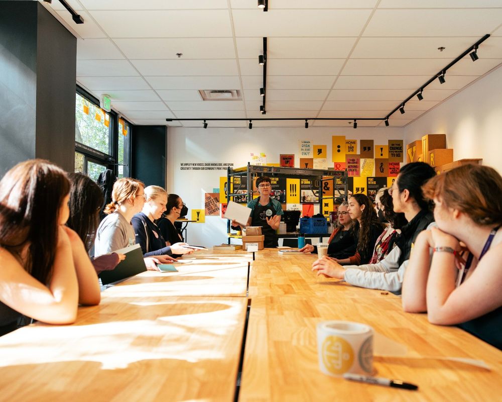 Students from the University of Washington Textual Studies 402: Book Arts course sit at a rectangular table while listening to a speaker at Partners in Print.