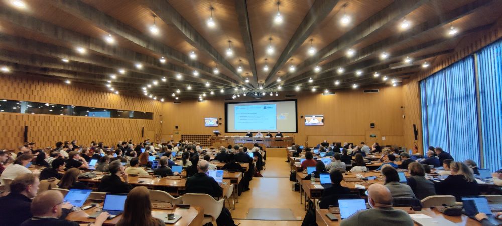 A spacious conference room with wooden panel walls and ceiling beams adorned with multiple hanging lights. The focus is on a central stage with a presentation screen displaying the topic "Cyber and Security Regulations" and logos of Software Heritage and UNESCO. Panellists are seated at a long table, with audience members filling the room, many using laptops at their desks. The atmosphere reflects an engaging and professional conference setting.

