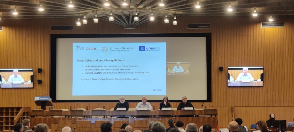 A conference room with wooden panel walls, featuring a panel discussion titled "Cyber and Security Regulations" on a central screen. The screen displays the Software Heritage and UNESCO logos, along with the names of panellists: Mike Milinkovich (Eclipse Foundation), Olivier Zendra (Inria), Carolina Lavelli (Internet of Trust), and moderator Simon Phipps (AlmaLinux OS Foundation). Panellists are seated at a long table below the screen, and side monitors show close-ups of the speakers. Audience members are visible in the foreground.