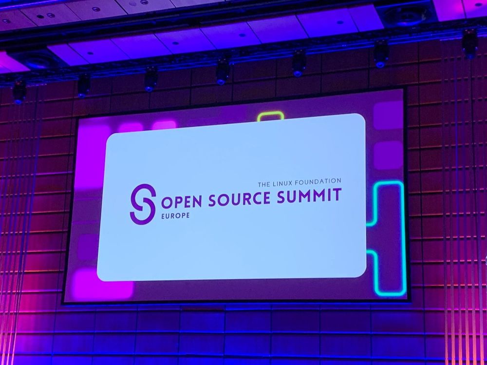 Photographie d'un écran de présentation lors de l'Open Source Summit Europe, organisé par la Linux Foundation. L'écran affiche le logo et le nom de l'événement 'Open Source Summit Europe' avec un fond violet et des motifs géométriques colorés. L'éclairage ambiant est également violet, créant une atmosphère moderne et technologique.