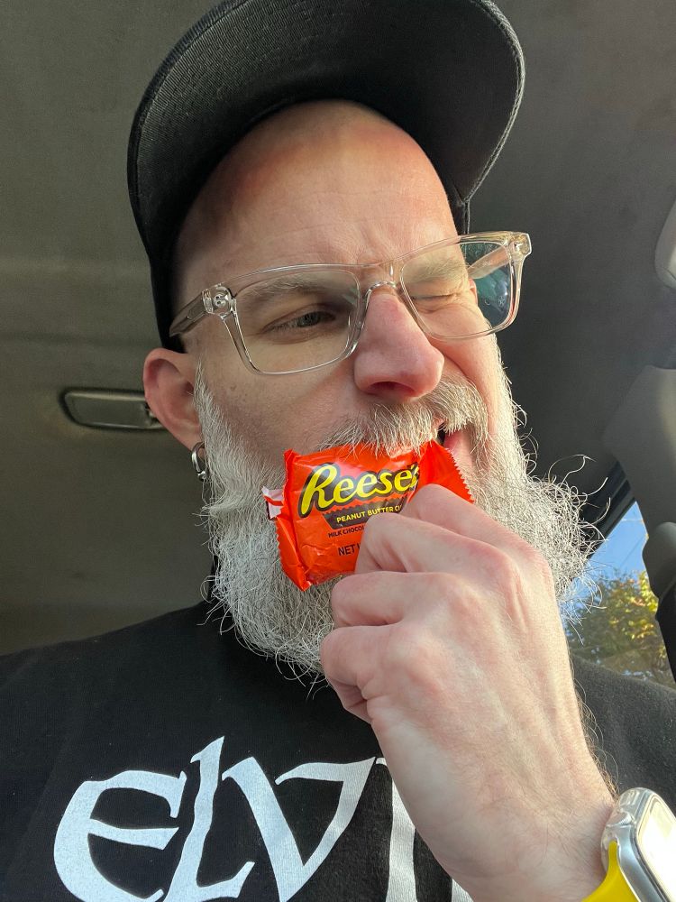 Bearded individual ravenously tearing into their favorite Halloween candy: Reese’s Peanut Butter Cups