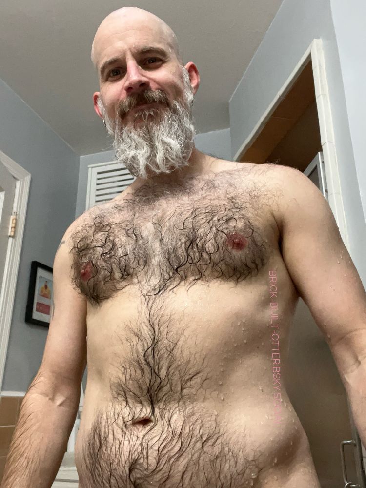 Bearded and hirsute individual standing in front of the shower, water droplets dot their body, chest and belly hair matted in thick curls and swirls to their skin.