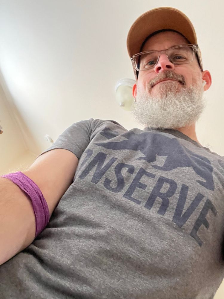 Bearded individual wearing glasses and a hat stares down at the camera, holding out an arm that was bandaged up after having blood drawn.  