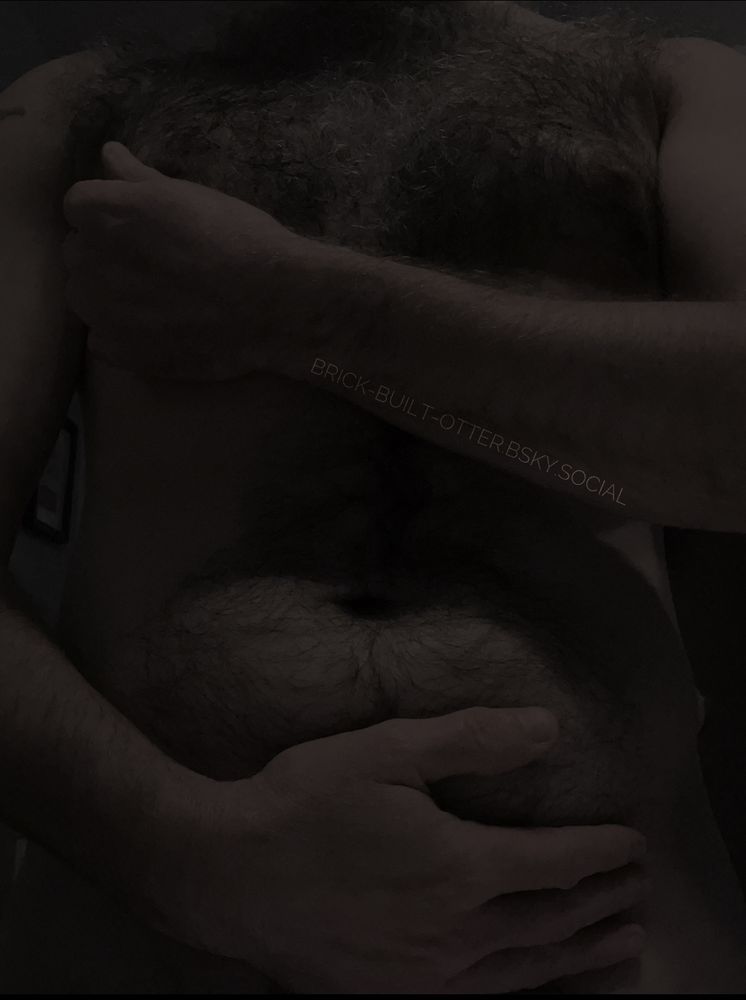 Same torso shot of a hirsute individual, one arm reaches across their curly haired chest to cup a tiddy while the other cradles a fuzzy belly, but this image is darker and more shadowed.
