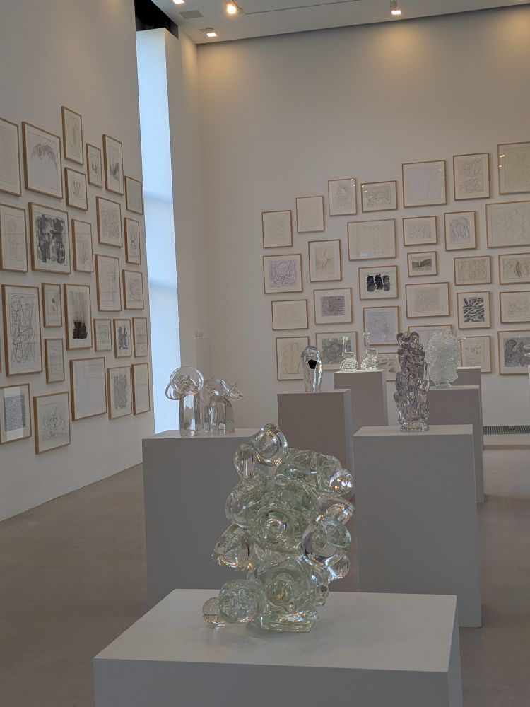 Eine Ausstellungshalle mit Glasskulpturen und vielen Zeichnungen an den Wänden