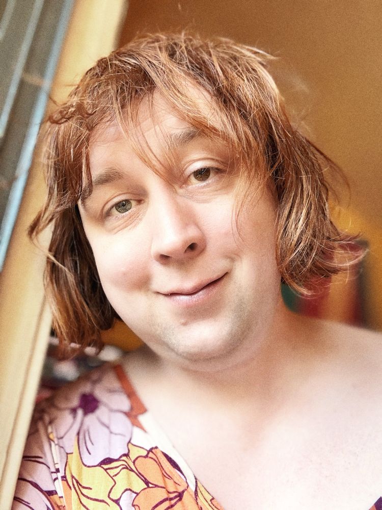 Photo of Lily from this week. Her hair is worn down in curls and her shirt is worn off one shoulder and it has pink yellow and orange flowers on it. She's not wearing glasses. 