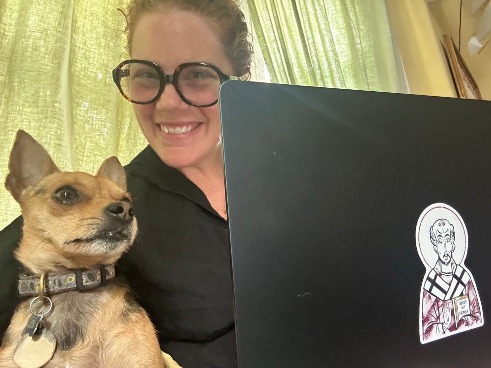 A tiny chihuahua supervises a lady pastor at a computer. 