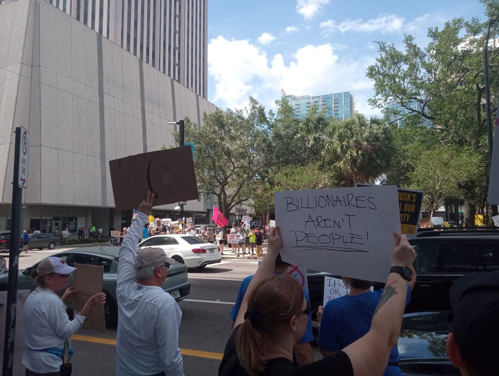 Woman holding poster with text "Billionaires aren't People" with crowd of protesters across the street.