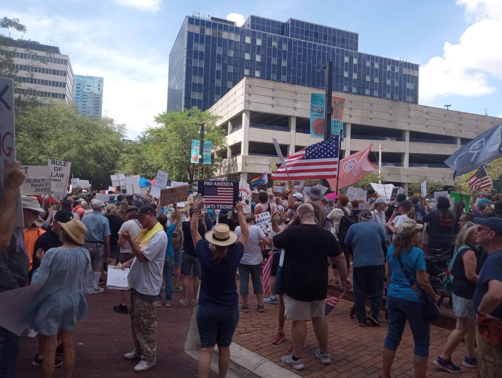 Back of crowd at protest, many holding signs, in downtown. 