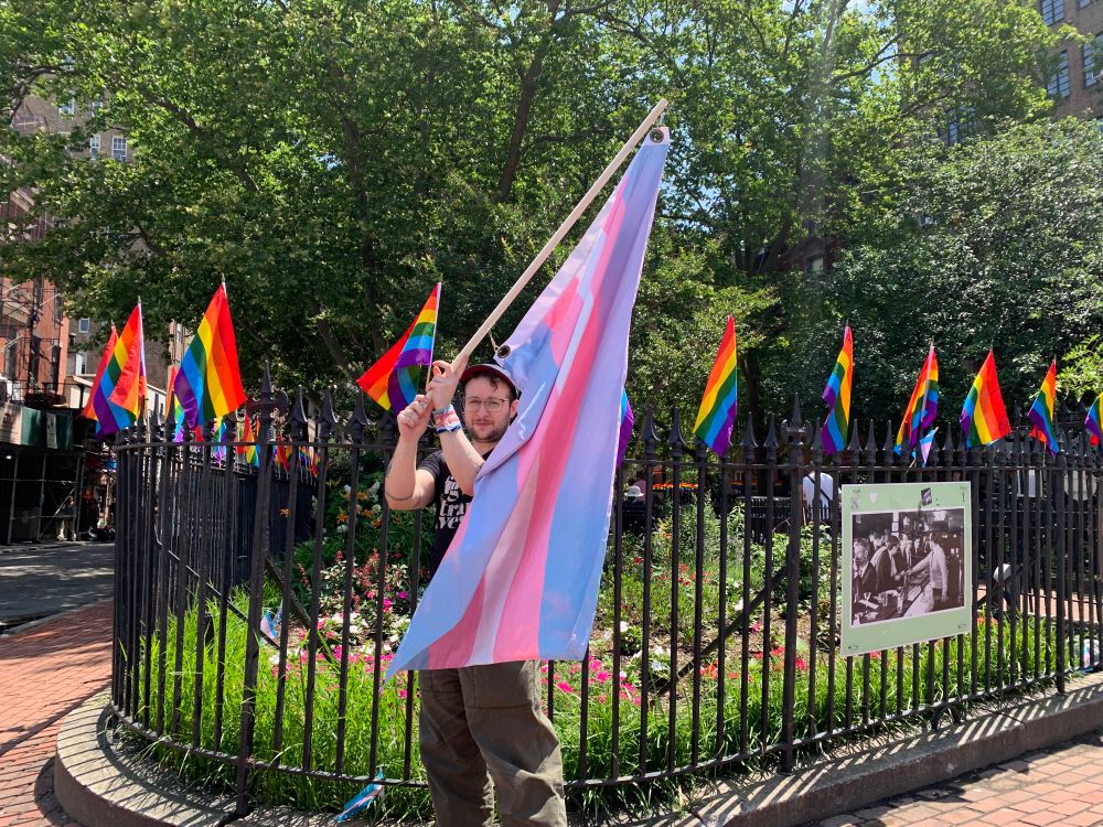 A white man holds a large trans flag just outside Stonewall National Monument