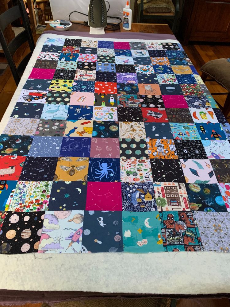 An assembled but unsewn quilt. The top is a cacophony of patterned squares, set over slightly larger cotton batting. The backing is not visible.