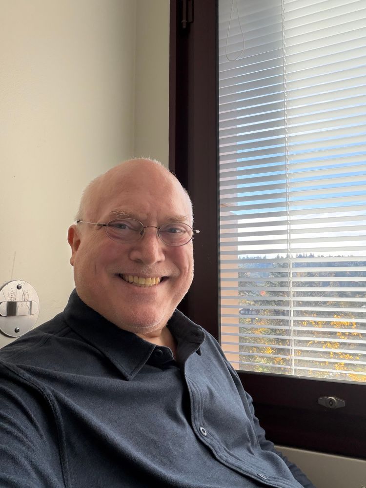 Photo of Glenn in buttoned shirt in front of window with slats and trees beyond