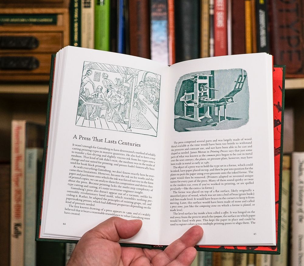 Hand holding open book in front of slightly out of focus bookshelves. The left and right book pages have illustrations at the top: Dance of Death (left); Common Press (right)