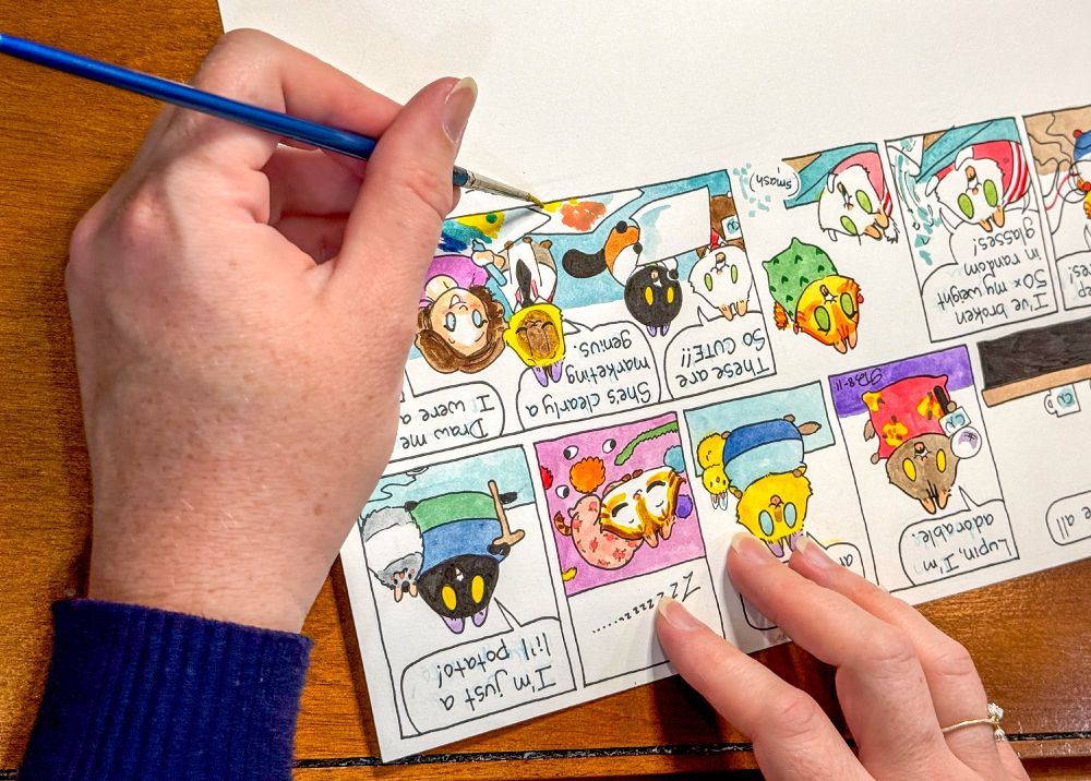 Hand with watercolor brush applying details to comic strip Breaking Cat News (Georgia Dunn). The strip is oriented upside down as the cartoonist steadies it with one hand and uses her other to paint.