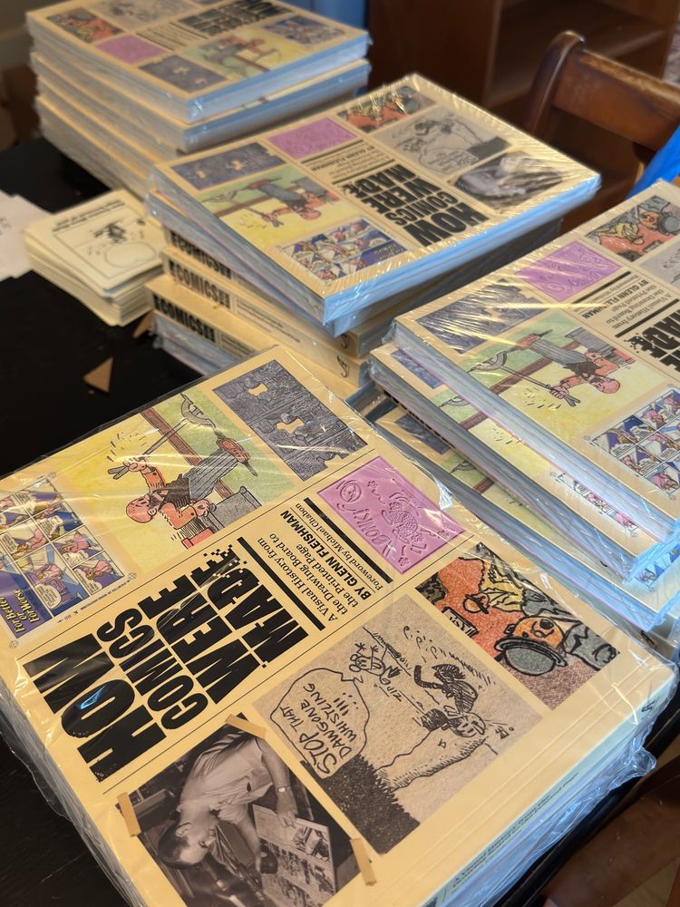 Piles of How Comics Were Made book: yellow covers, wrapped in plastic, with the title and images of cartoonists and printing artifacts all around the title.