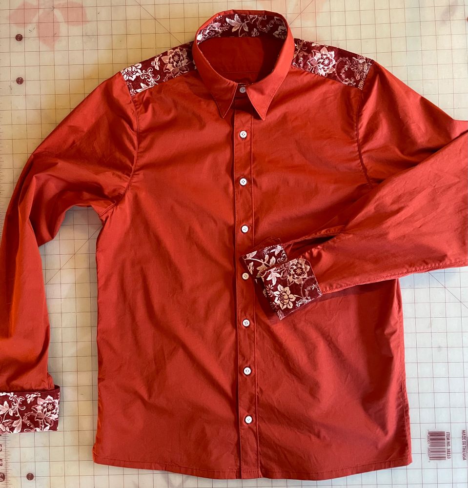A button front shirt with a pintuck pleated front placket, narrow winged collar, and French cuffs. It is made of rust red cotton shirt with floral (roses and bees in white on a red background) accents at the yoke, inner collar band, and cuffs.