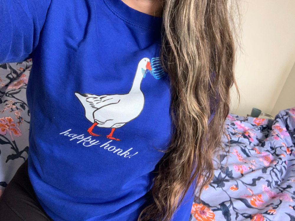 A blue sweatshirt with a picture of a goose holding a menorah, underneath the goose it says “happy honk!”
