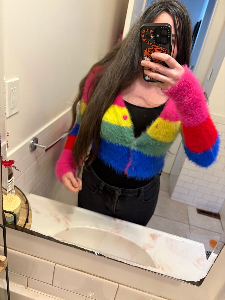 A picture in a mirror of a woman in a fuzzy rainbow sweater with hair down to her waist
