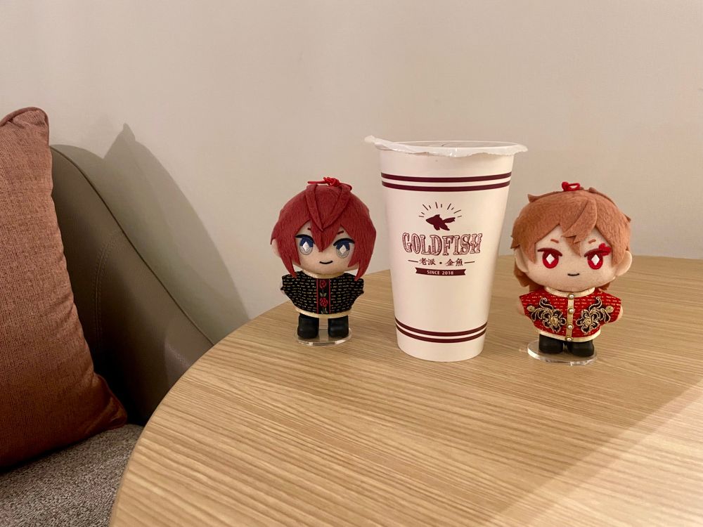 写真。ぬい撮り。ホテルの木製の丸テーブルの上にリドルくんのぬいぐるみとエースのぬいぐるみがタピオカの紙コップを挟んで立っている。白い紙コップには赤茶色で「GOLD FISH ー老派・金魚ー」の文字と金魚のシルエットがプリントされている。ぬいは二人とも漢服風のぬい服を着ている。リドぬいは黒い服で胸にバラの模様がある。エスぬいは赤い服で胸に黒い花の刺繍のような模様がある。