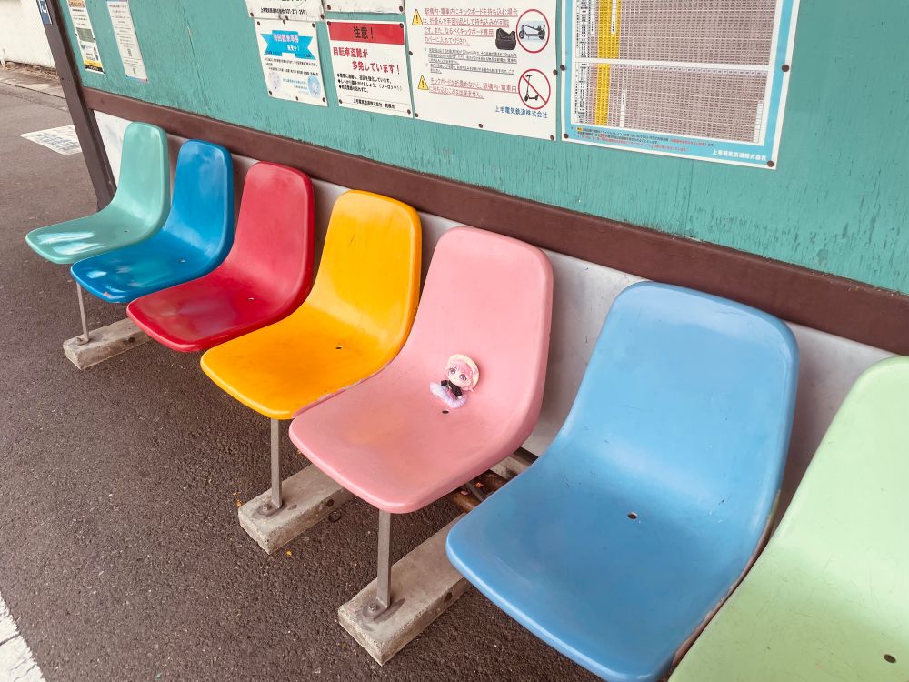 写真。上毛電気鉄道三俣駅のホーム。緑青赤黄色ピンク…とカラフルな椅子が並んでいる。ピンクの椅子の上にユイナぬい。